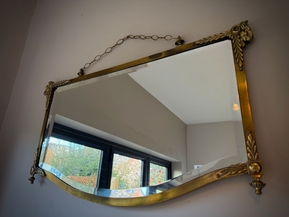 Horizontal wall mirror featuring a brass frame with sculpted rams heads on the top corners, leaf motifs, a curved lower edge, and an overhead hanging chain. View II