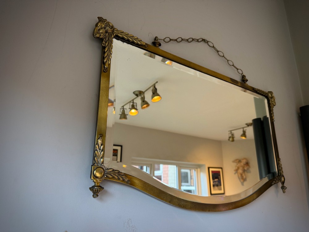 Horizontal wall mirror featuring a brass frame with sculpted rams heads on the top corners, leaf motifs, a curved lower edge, and an overhead hanging chain. View III