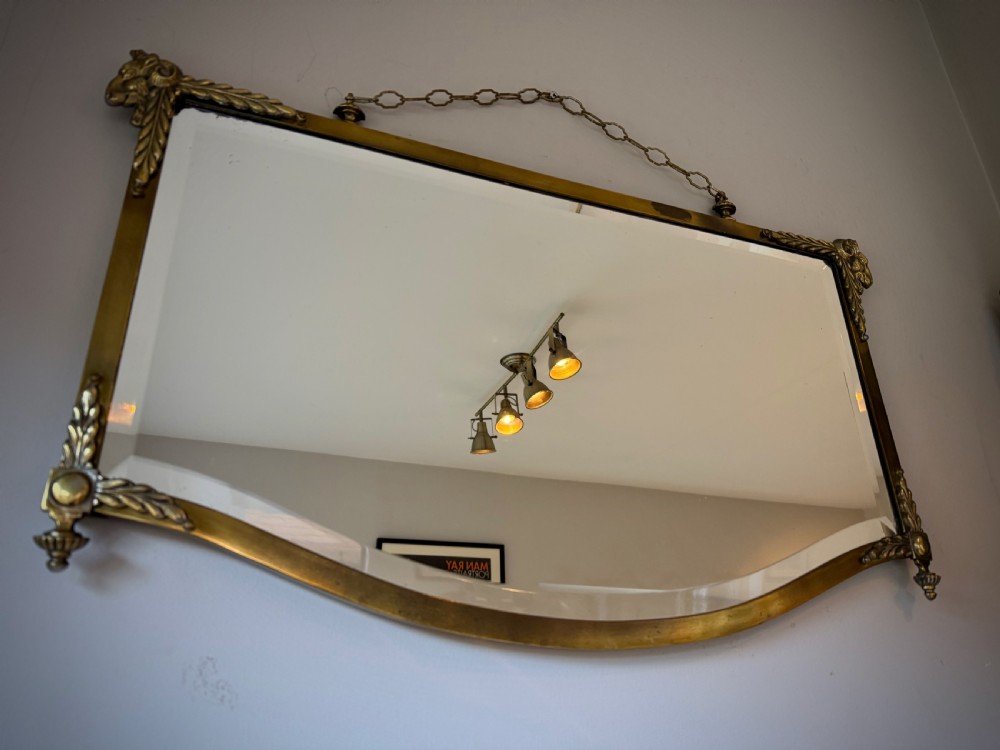 Horizontal wall mirror featuring a brass frame with sculpted rams heads on the top corners, leaf motifs, a curved lower edge, and an overhead hanging chain. View IV
