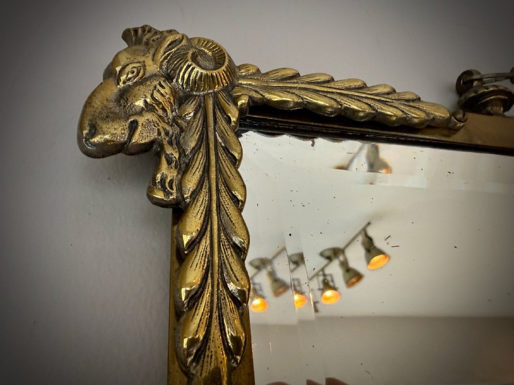 Horizontal wall mirror featuring a brass frame with sculpted rams heads on the top corners, leaf motifs, a curved lower edge, and an overhead hanging chain. View V
