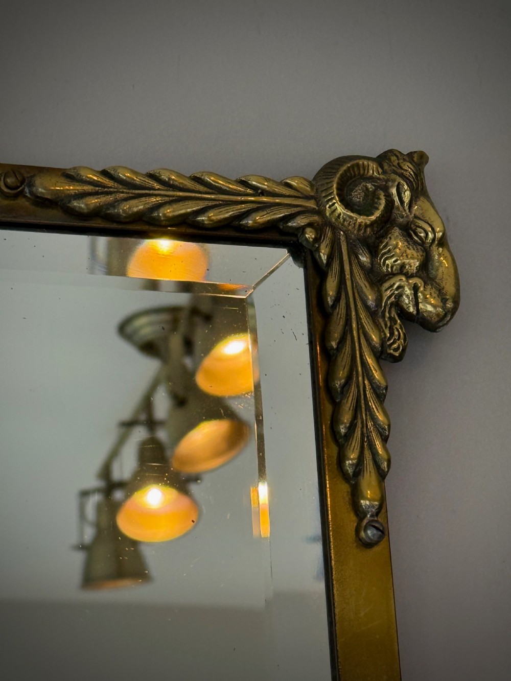 Horizontal wall mirror featuring a brass frame with sculpted rams heads on the top corners, leaf motifs, a curved lower edge, and an overhead hanging chain. View VI