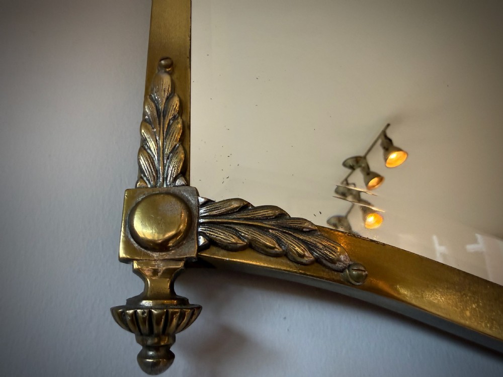 Horizontal wall mirror featuring a brass frame with sculpted rams heads on the top corners, leaf motifs, a curved lower edge, and an overhead hanging chain. View VII
