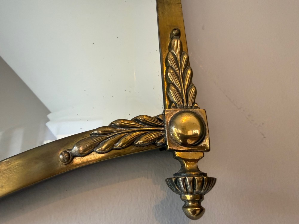 Horizontal wall mirror featuring a brass frame with sculpted rams heads on the top corners, leaf motifs, a curved lower edge, and an overhead hanging chain. View VIII