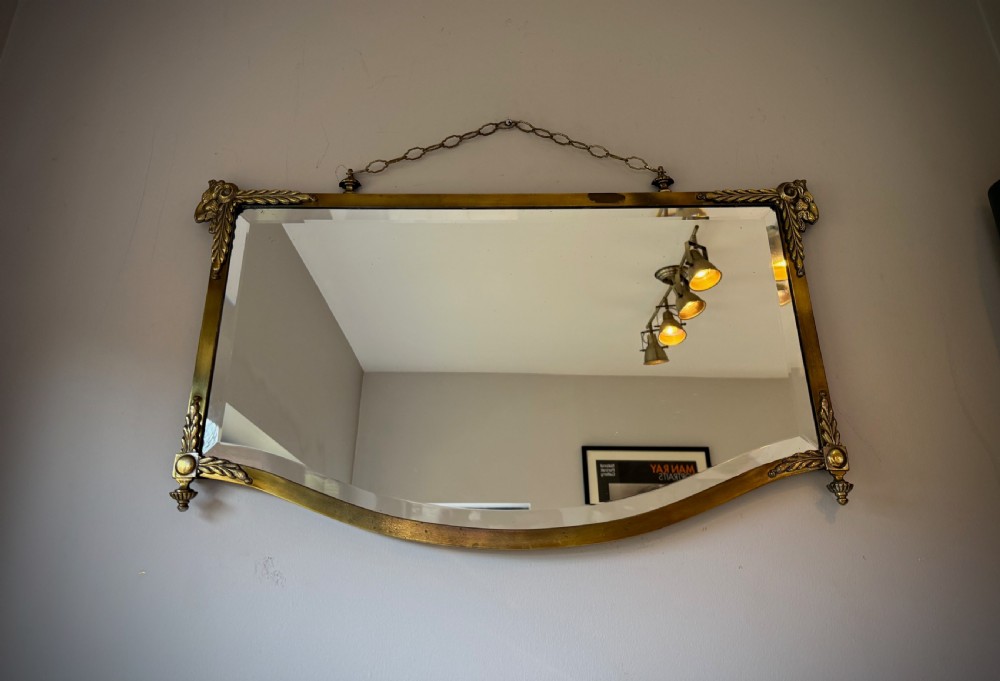 Horizontal wall mirror featuring a brass frame with sculpted rams heads on the top corners, leaf motifs, a curved lower edge, and an overhead hanging chain. View X