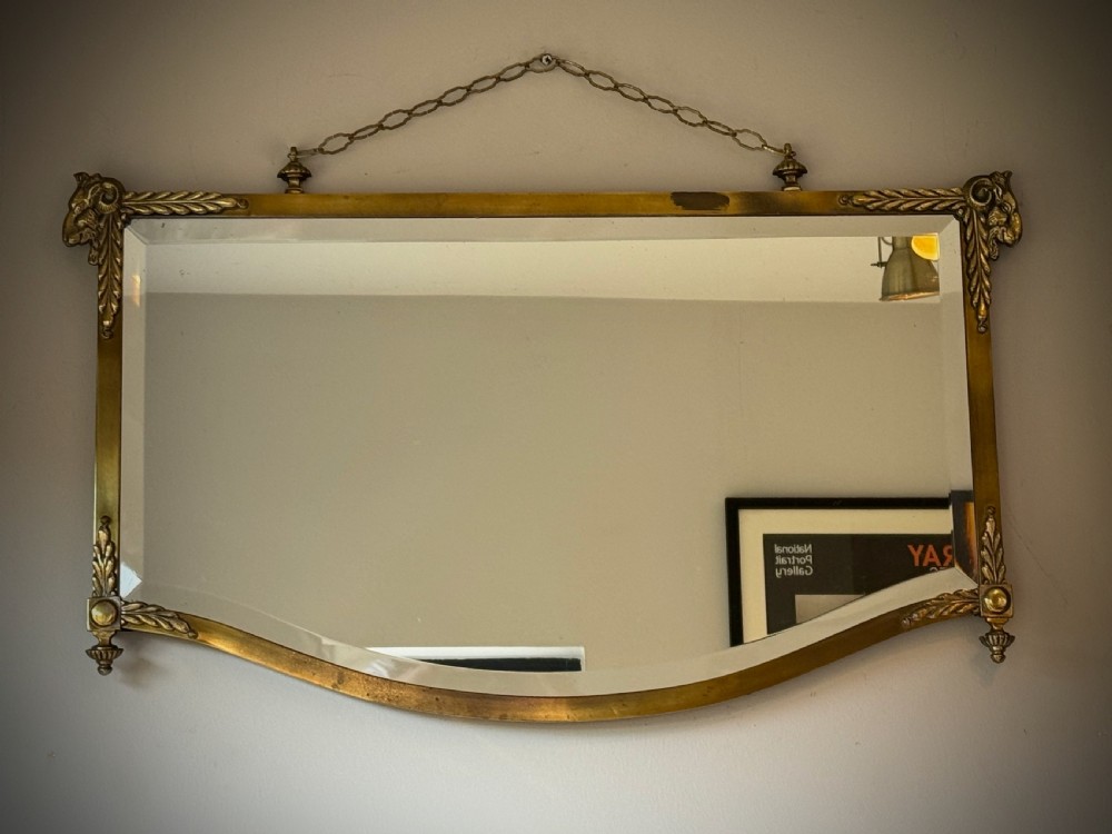 Horizontal wall mirror featuring a brass frame with sculpted rams heads on the top corners, leaf motifs, a curved lower edge, and an overhead hanging chain. View XI