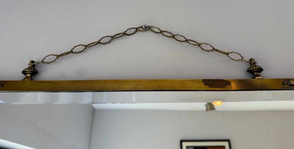 Horizontal wall mirror featuring a brass frame with sculpted rams heads on the top corners, leaf motifs, a curved lower edge, and an overhead hanging chain. View XII