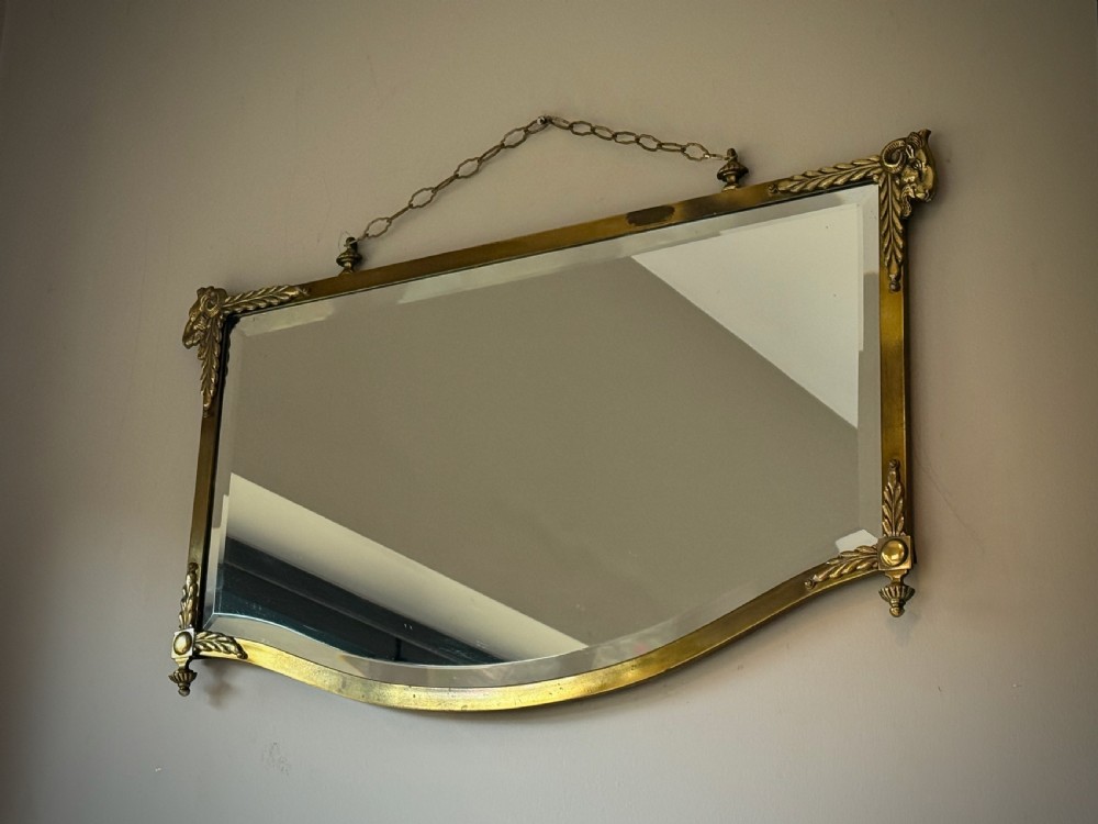 Horizontal wall mirror featuring a brass frame with sculpted rams heads on the top corners, leaf motifs, a curved lower edge, and an overhead hanging chain. View XIII