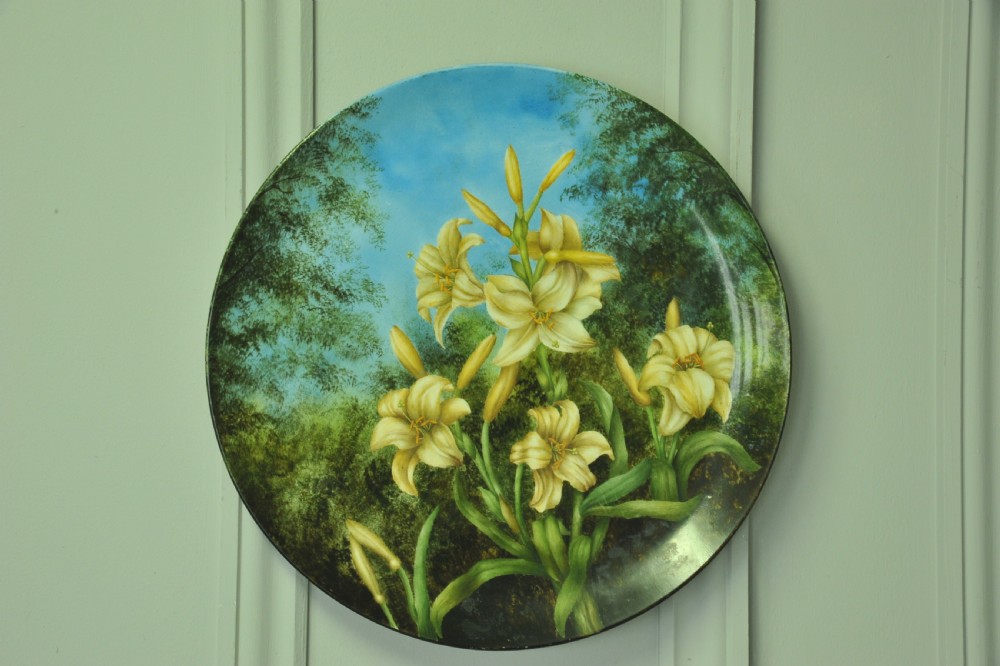 A circular hand-painted porcelain decorative plate displaying yellow lilies against foliage and a blue sky.