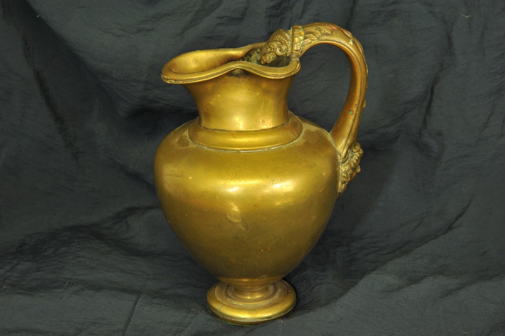 An antique brass oinochoe wine jug featuring a trefoil-shaped spout, a bulbous body on a pedestal foot, and a high loop handle adorned with a carved ram head motif.
