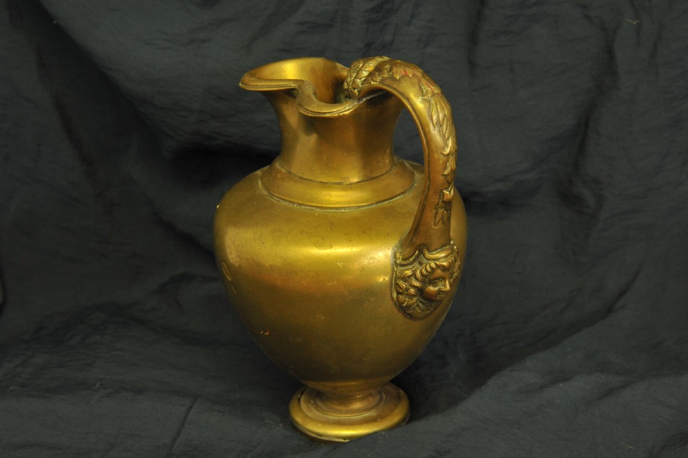 An antique brass oinochoe wine jug featuring a trefoil-shaped spout, a bulbous body on a pedestal foot, and a high loop handle adorned with a carved ram head motif. View II