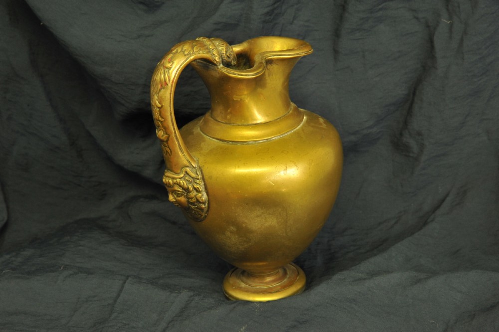 An antique brass oinochoe wine jug featuring a trefoil-shaped spout, a bulbous body on a pedestal foot, and a high loop handle adorned with a carved ram head motif. View III