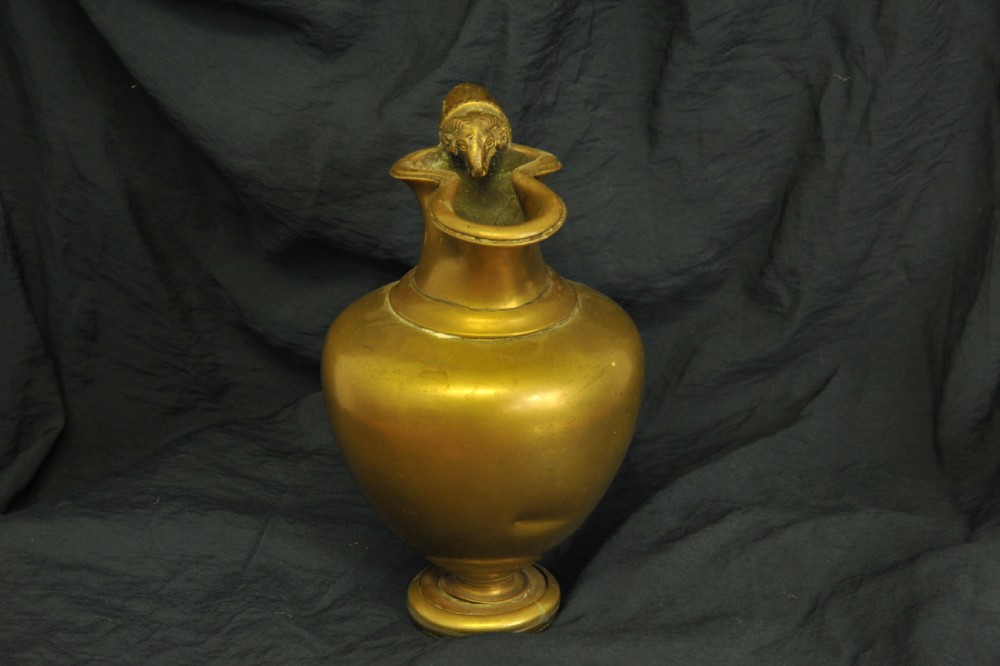 An antique brass oinochoe wine jug featuring a trefoil-shaped spout, a bulbous body on a pedestal foot, and a high loop handle adorned with a carved ram head motif. View IV
