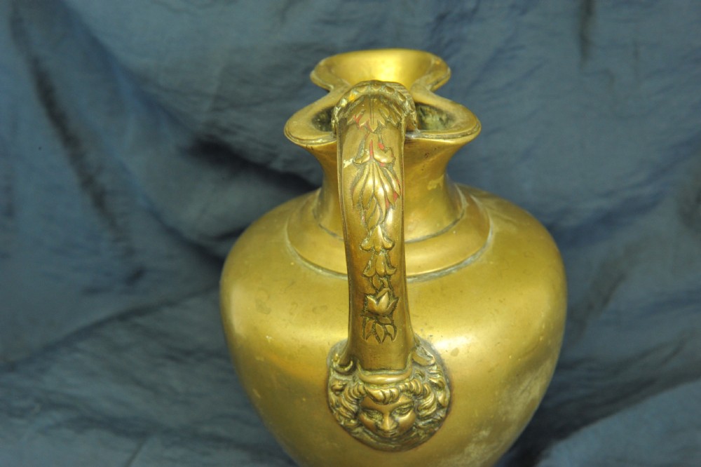 An antique brass oinochoe wine jug featuring a trefoil-shaped spout, a bulbous body on a pedestal foot, and a high loop handle adorned with a carved ram head motif. View V