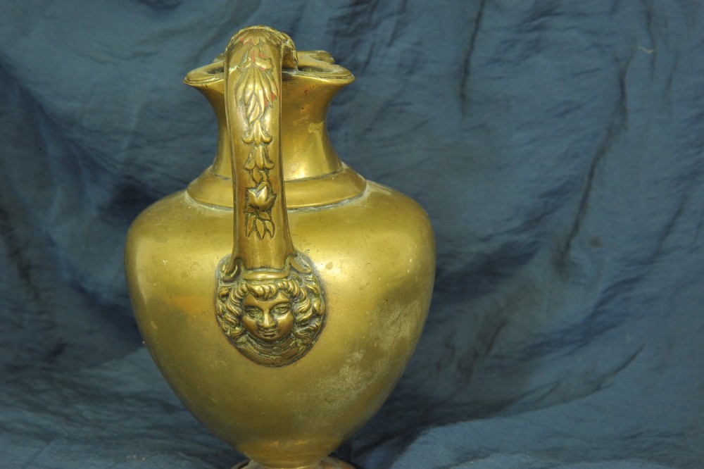 An antique brass oinochoe wine jug featuring a trefoil-shaped spout, a bulbous body on a pedestal foot, and a high loop handle adorned with a carved ram head motif. View VI