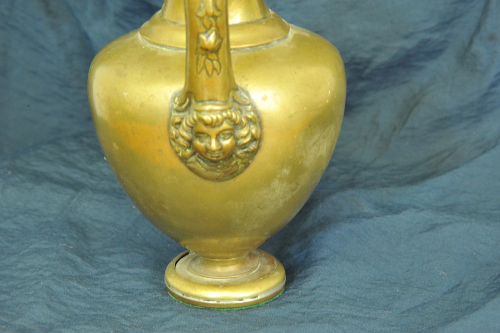 An antique brass oinochoe wine jug featuring a trefoil-shaped spout, a bulbous body on a pedestal foot, and a high loop handle adorned with a carved ram head motif. View VII