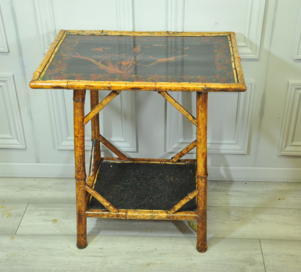 A late Victorian two-tier bamboo side table featuring a dark lacquered top panel decorated with botanical flora and birds.