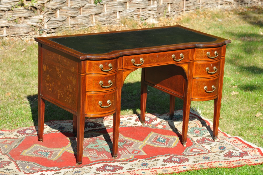 An antique Edwardian inlaid mahogany writing desk featuring a dark leather inset top, seven drawers with brass handles, and floral marquetry side panels.