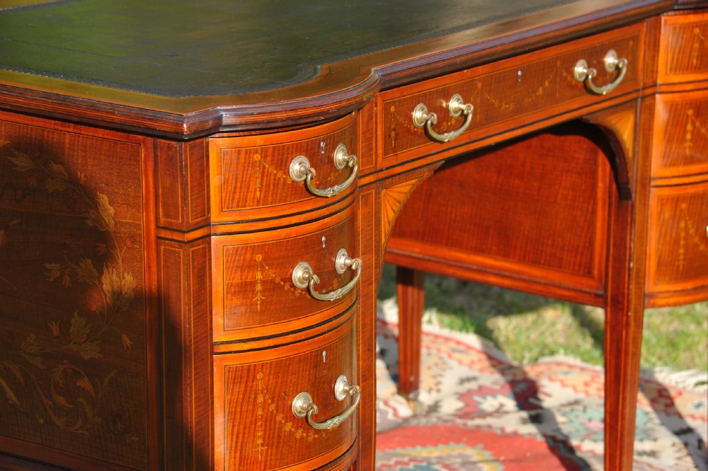 An antique Edwardian inlaid mahogany writing desk featuring a dark leather inset top, seven drawers with brass handles, and floral marquetry side panels. View V