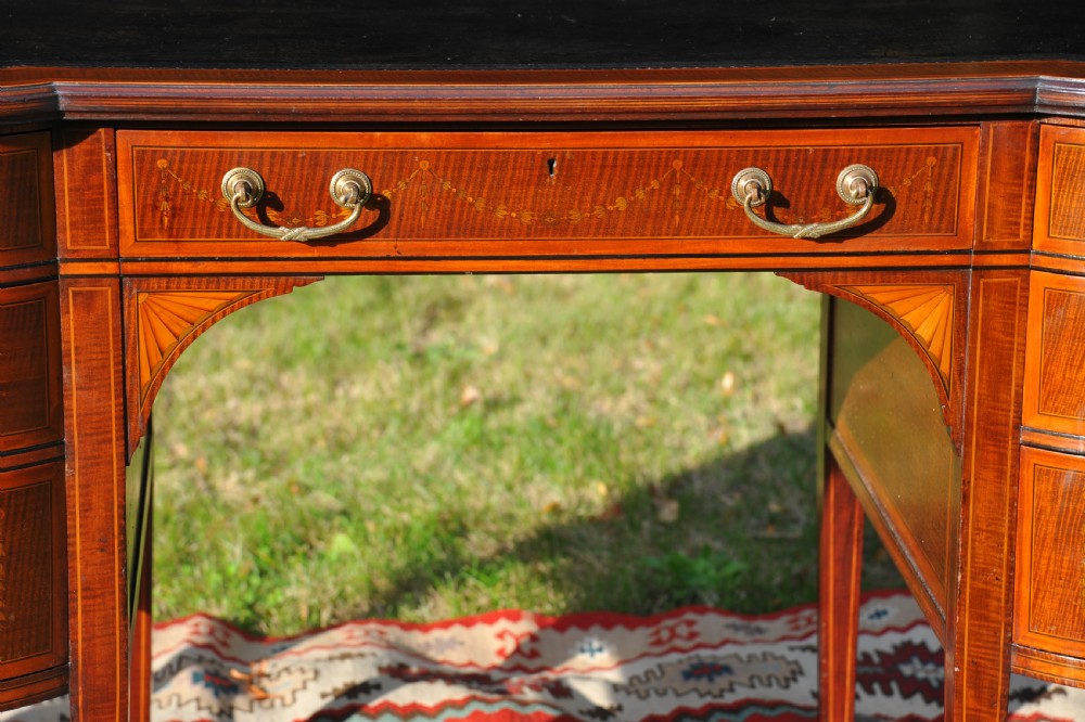 An antique Edwardian inlaid mahogany writing desk featuring a dark leather inset top, seven drawers with brass handles, and floral marquetry side panels. View VI