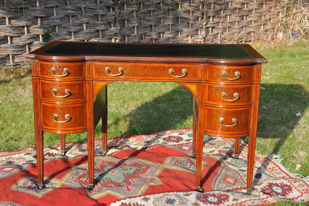 An antique Edwardian inlaid mahogany writing desk featuring a dark leather inset top, seven drawers with brass handles, and floral marquetry side panels. View X