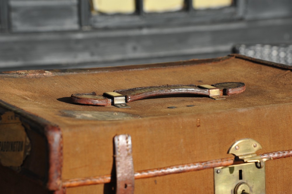 A square antique tan canvas travel case featuring stitched brown leather edging, a top carrying handle, brass rivets, and an authentic Paddington railway paper label. View V