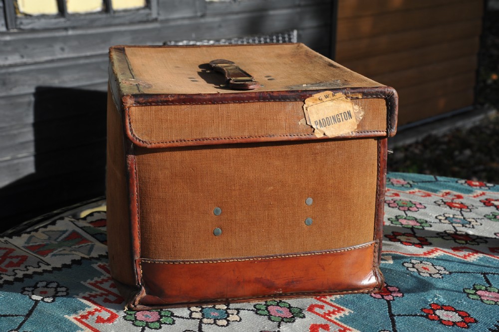A square antique tan canvas travel case featuring stitched brown leather edging, a top carrying handle, brass rivets, and an authentic Paddington railway paper label. View VII