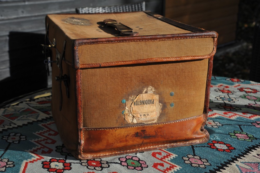 A square antique tan canvas travel case featuring stitched brown leather edging, a top carrying handle, brass rivets, and an authentic Paddington railway paper label.