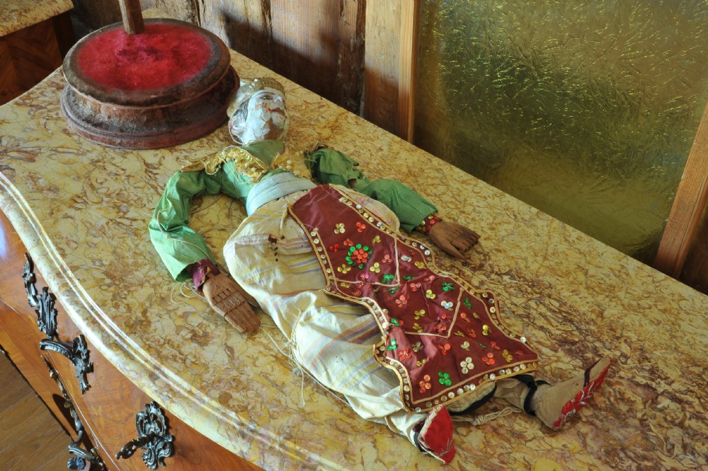 An antique carved wooden marionette puppet wearing a green top and embellished red and white skirt, mounted on a circular base. View V