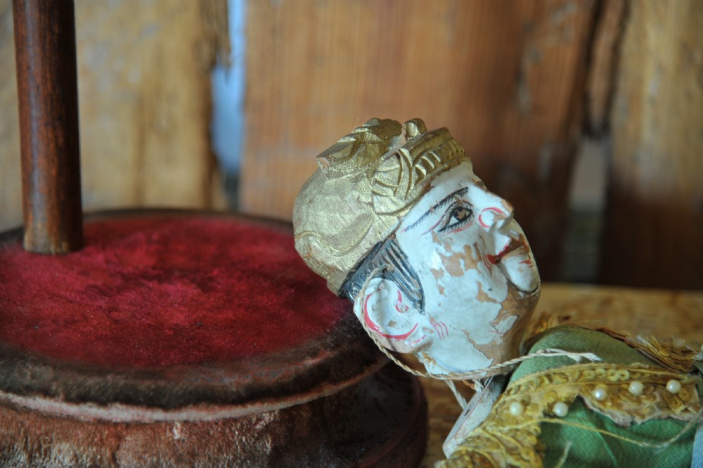 An antique carved wooden marionette puppet wearing a green top and embellished red and white skirt, mounted on a circular base. View VII