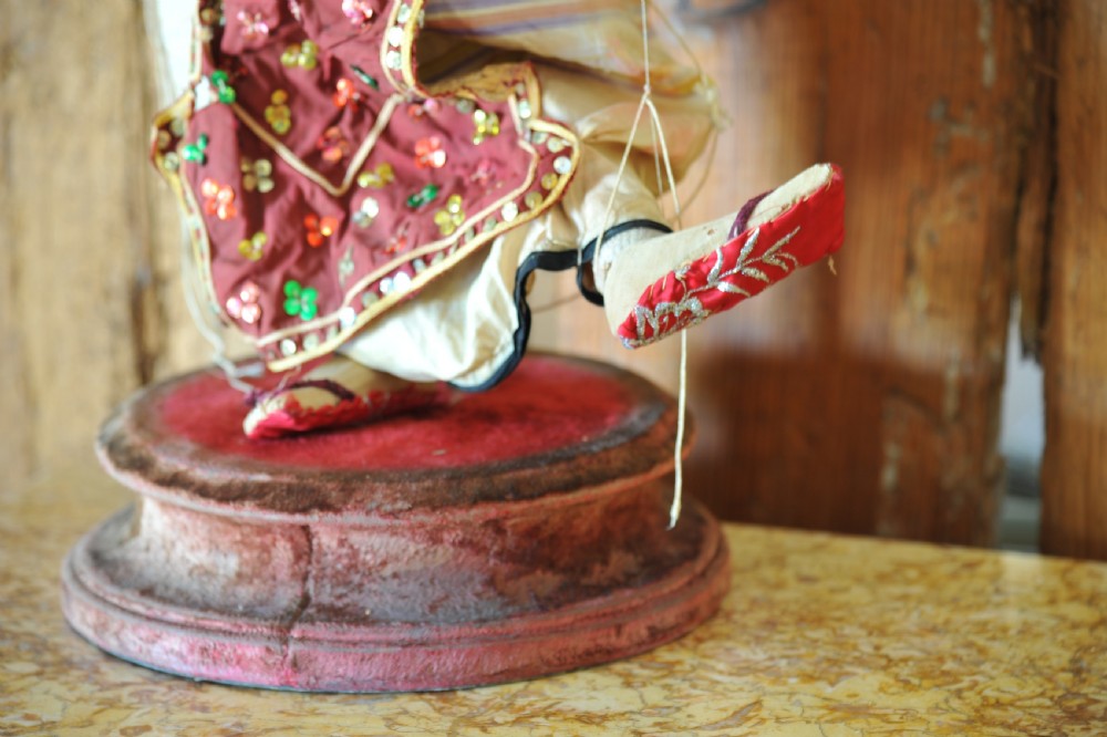 An antique carved wooden marionette puppet wearing a green top and embellished red and white skirt, mounted on a circular base. View XIII