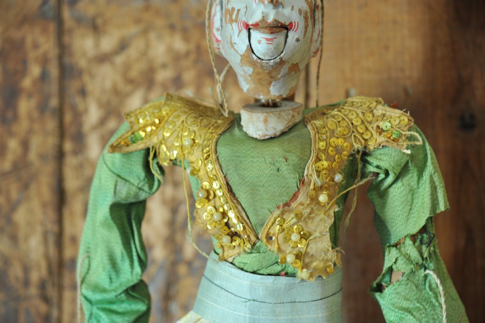 An antique carved wooden marionette puppet wearing a green top and embellished red and white skirt, mounted on a circular base. View XIV