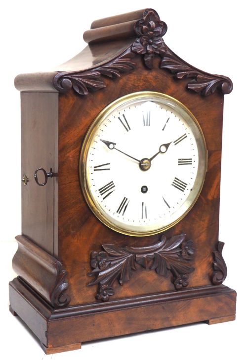 fig 1. An antique Victorian flame mahogany bracket clock featuring carved leaf decoration, a round cream dial with Roman numerals, and a hinged brass bezel.