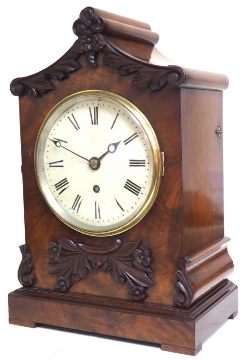 fig 2. An antique Victorian flame mahogany bracket clock featuring carved leaf decoration, a round cream dial with Roman numerals, and a hinged brass bezel.