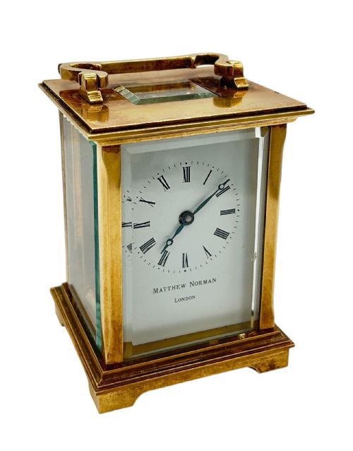 fig 1. A rectangular brass Matthew Norman carriage clock with bevelled glass sides, a top carrying handle, and a white enamel dial displaying Roman numerals.