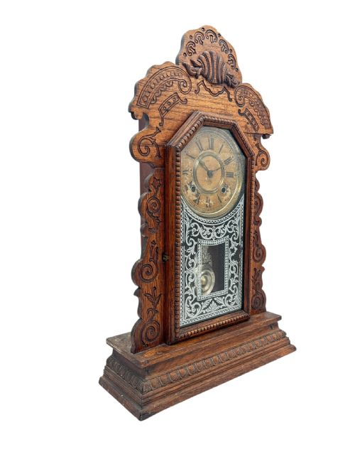 fig 1. An antique Victorian Ansonia gingerbread mantel clock crafted from carved oak with a round dial, brass pendulum, and stencilled glass door.