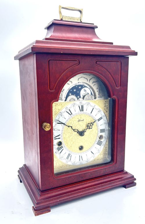 fig 2. A wooden bracket clock featuring a reddish mahogany case, brass carrying handle, and a decorative dial with moon phase indicator.