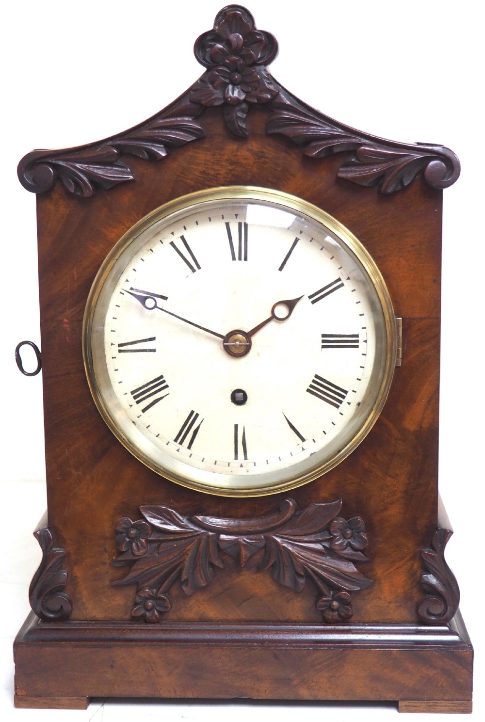 An antique Victorian flame mahogany bracket clock featuring carved leaf decoration, a round cream dial with Roman numerals, and a hinged brass bezel.