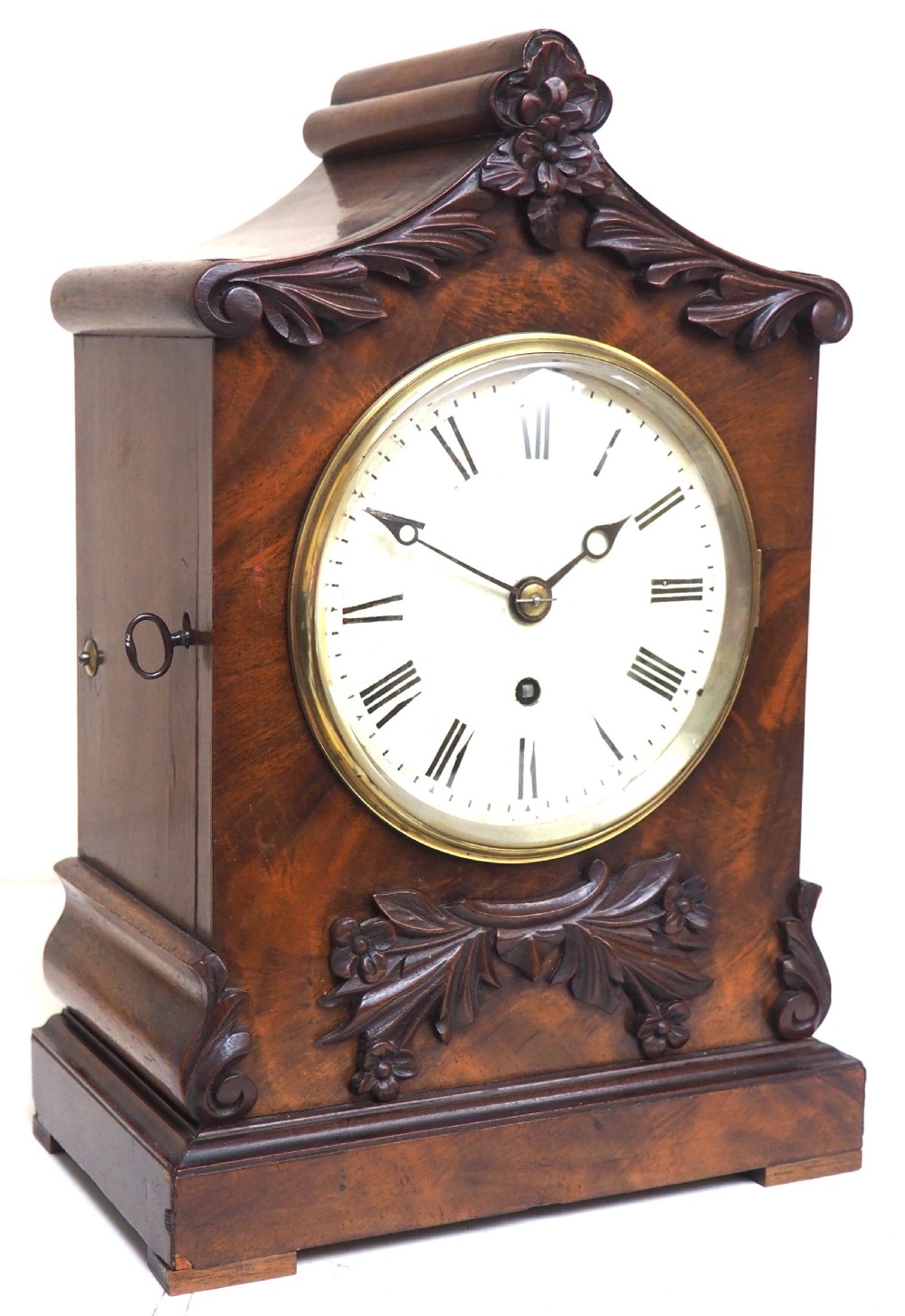 An antique Victorian flame mahogany bracket clock featuring carved leaf decoration, a round cream dial with Roman numerals, and a hinged brass bezel. View II