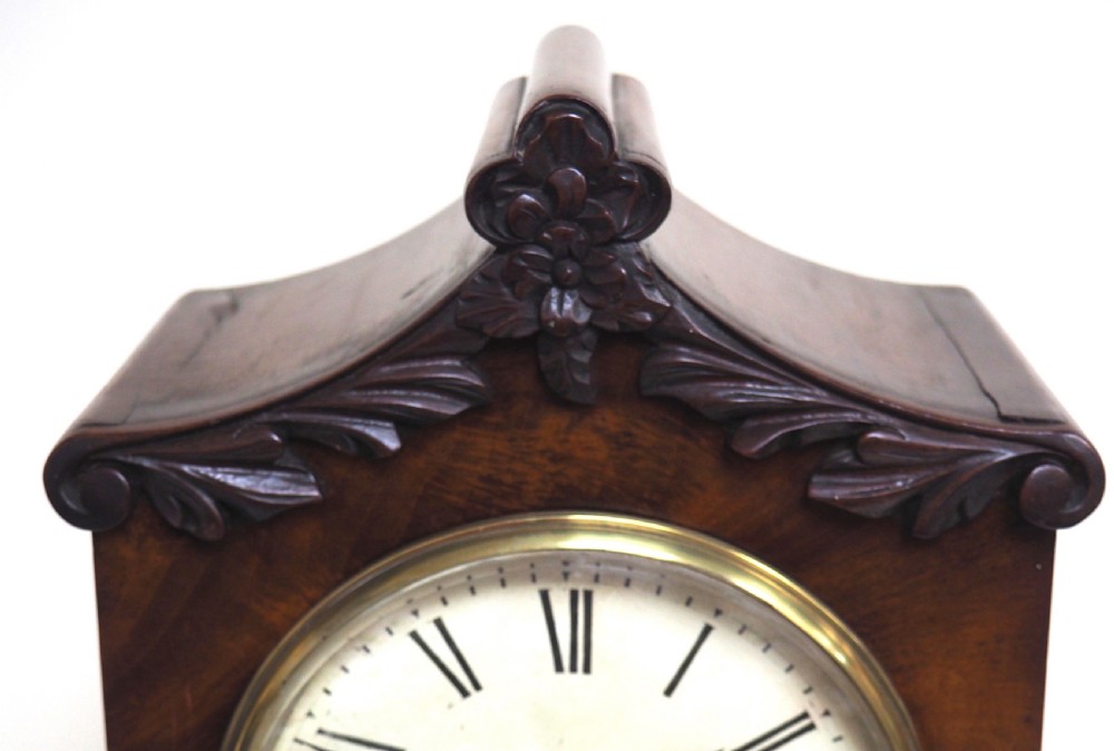An antique Victorian flame mahogany bracket clock featuring carved leaf decoration, a round cream dial with Roman numerals, and a hinged brass bezel. View IV