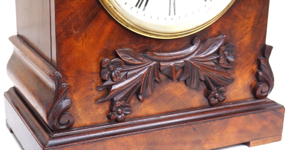 An antique Victorian flame mahogany bracket clock featuring carved leaf decoration, a round cream dial with Roman numerals, and a hinged brass bezel. View V