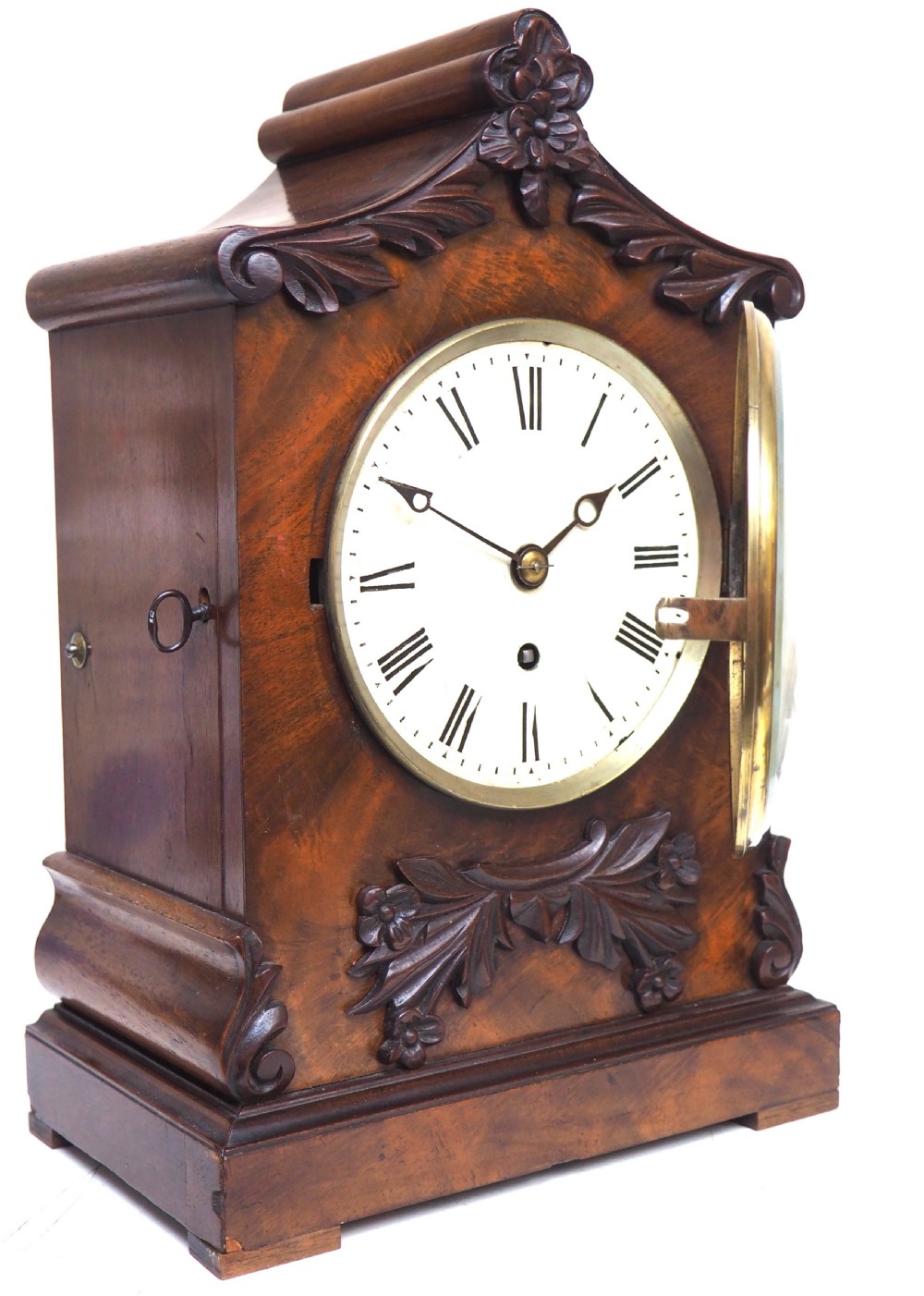 An antique Victorian flame mahogany bracket clock featuring carved leaf decoration, a round cream dial with Roman numerals, and a hinged brass bezel. View VI
