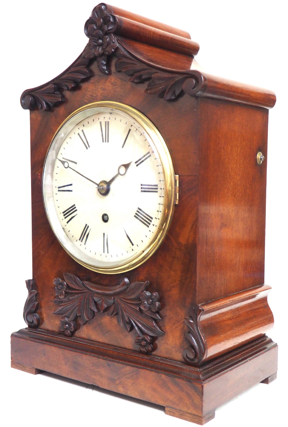 An antique Victorian flame mahogany bracket clock featuring carved leaf decoration, a round cream dial with Roman numerals, and a hinged brass bezel. View X