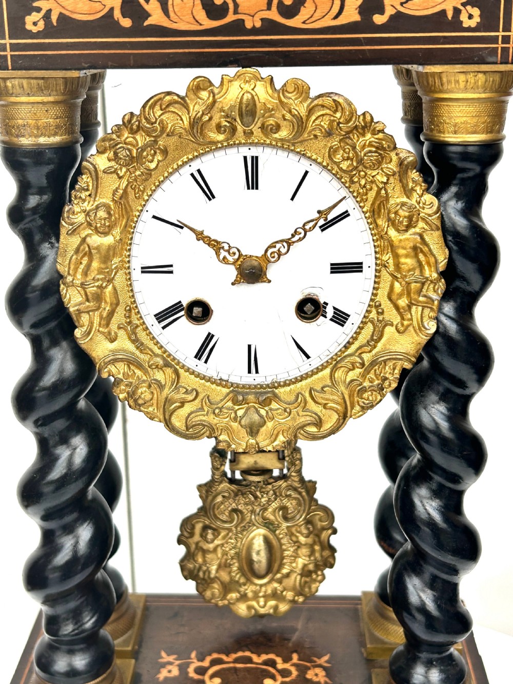 An antique French Empire ebonised and marquetry portico clock featuring four barley twist columns, gilt bronze capitals, an ornate ormolu framed enamel dial, and a decorative pendulum. View VI