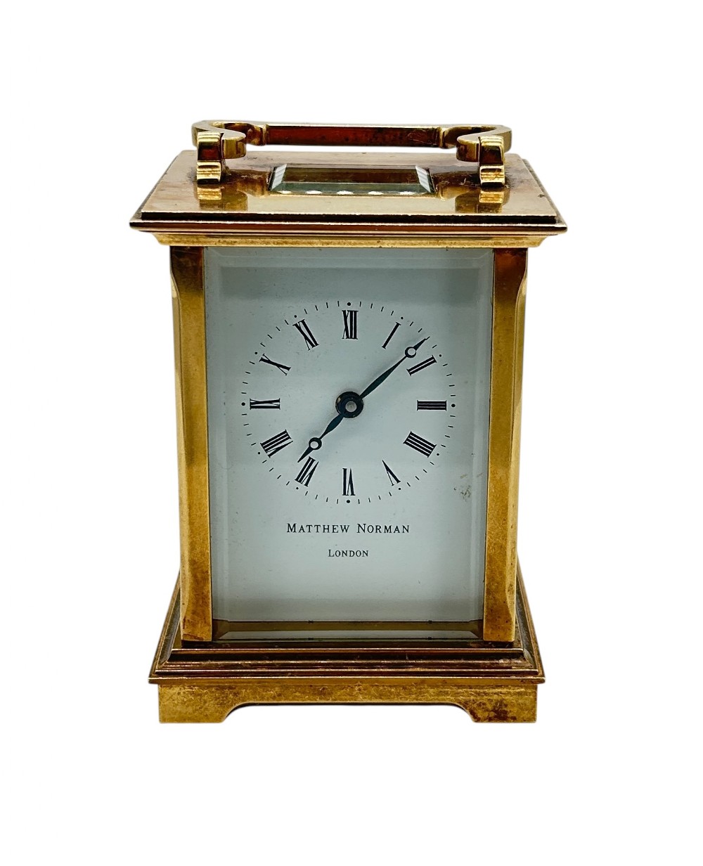 A rectangular brass Matthew Norman carriage clock with bevelled glass sides, a top carrying handle, and a white enamel dial displaying Roman numerals.