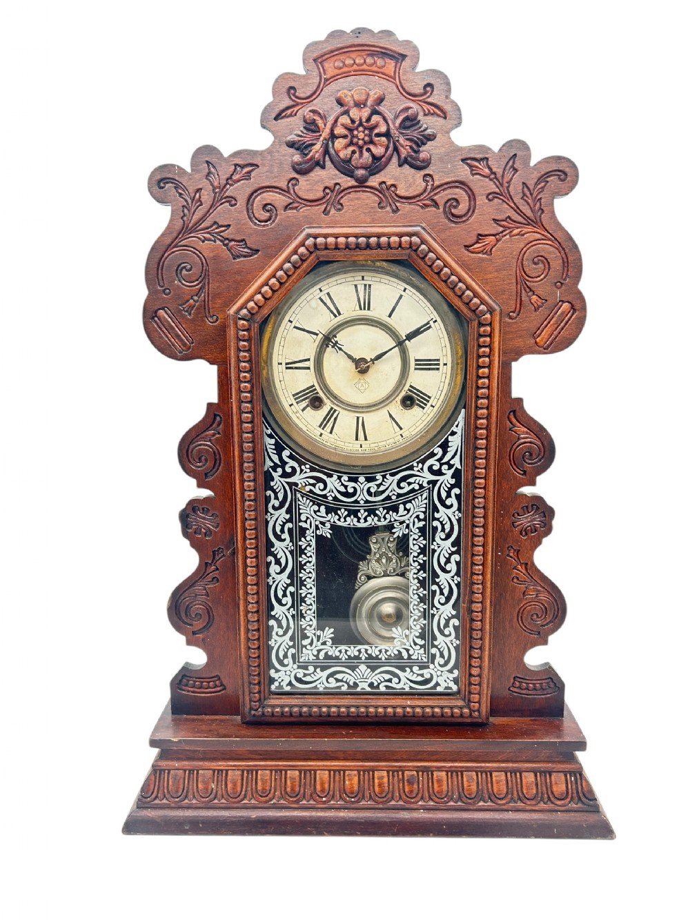 A late 19th-century Ansonia gingerbread mantel clock crafted from dark carved oak with a stencilled glass pendulum door and Roman numeral dial.