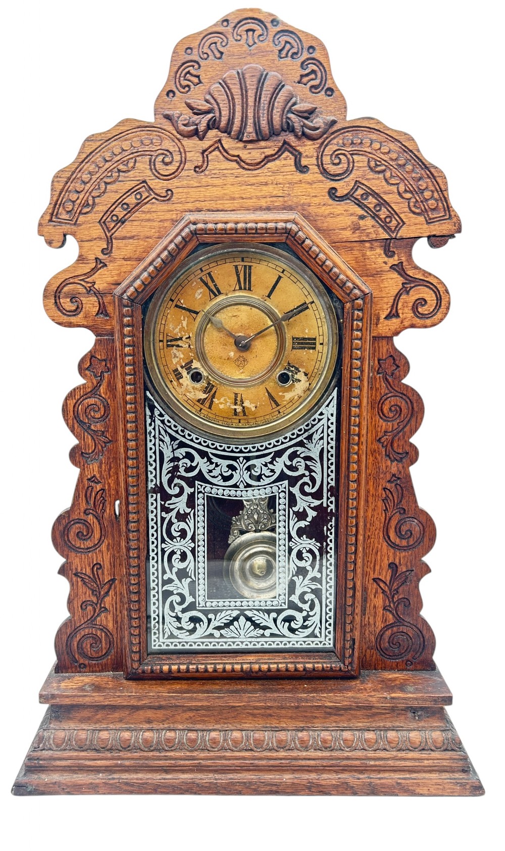 An antique Victorian Ansonia gingerbread mantel clock crafted from carved oak with a round dial, brass pendulum, and stencilled glass door.