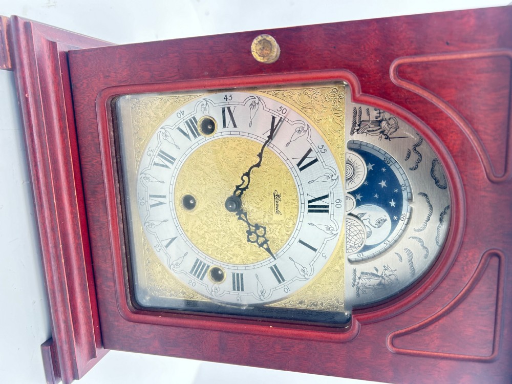 A wooden bracket clock featuring a reddish mahogany case, brass carrying handle, and a decorative dial with moon phase indicator. View IV