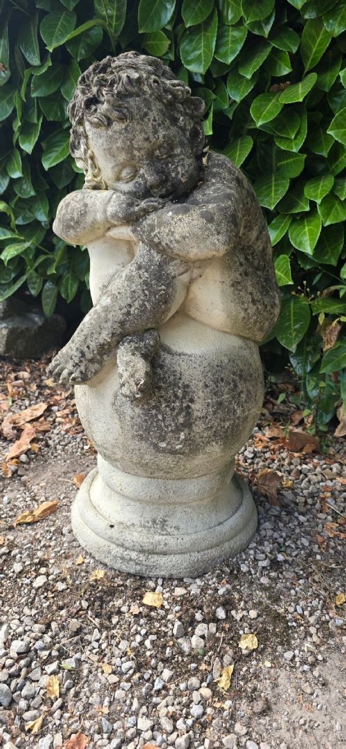 fig 1. A weathered grey cast stone statue of a sleeping cherub resting its head on crossed arms atop a spherical base and moulded pedestal.