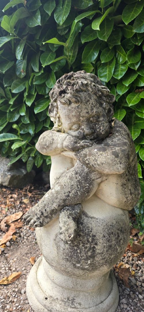 A weathered grey cast stone statue of a sleeping cherub resting its head on crossed arms atop a spherical base and moulded pedestal. View III