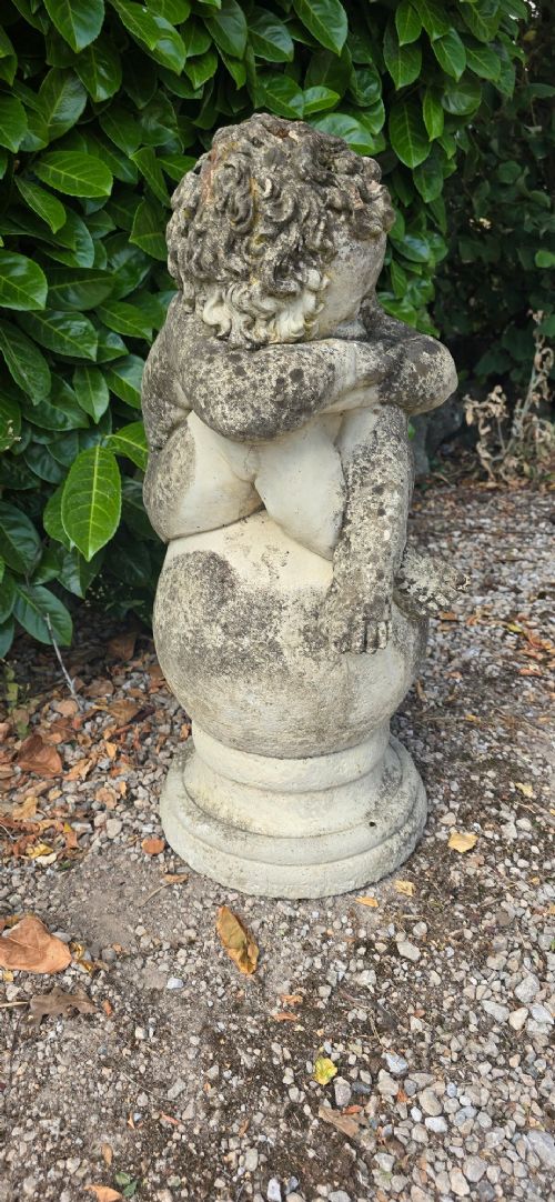 A weathered grey cast stone statue of a sleeping cherub resting its head on crossed arms atop a spherical base and moulded pedestal. View IV